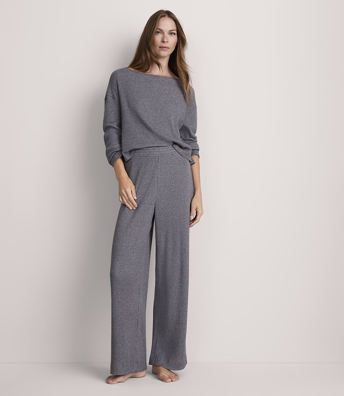 A woman stands in a grey ribbed loungewear set against a plain, light grey background.