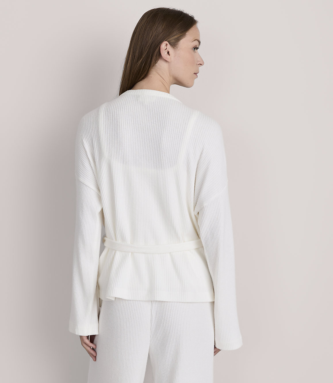 A woman in a white waffle-knit robe and pants stands against a plain, light gray background.