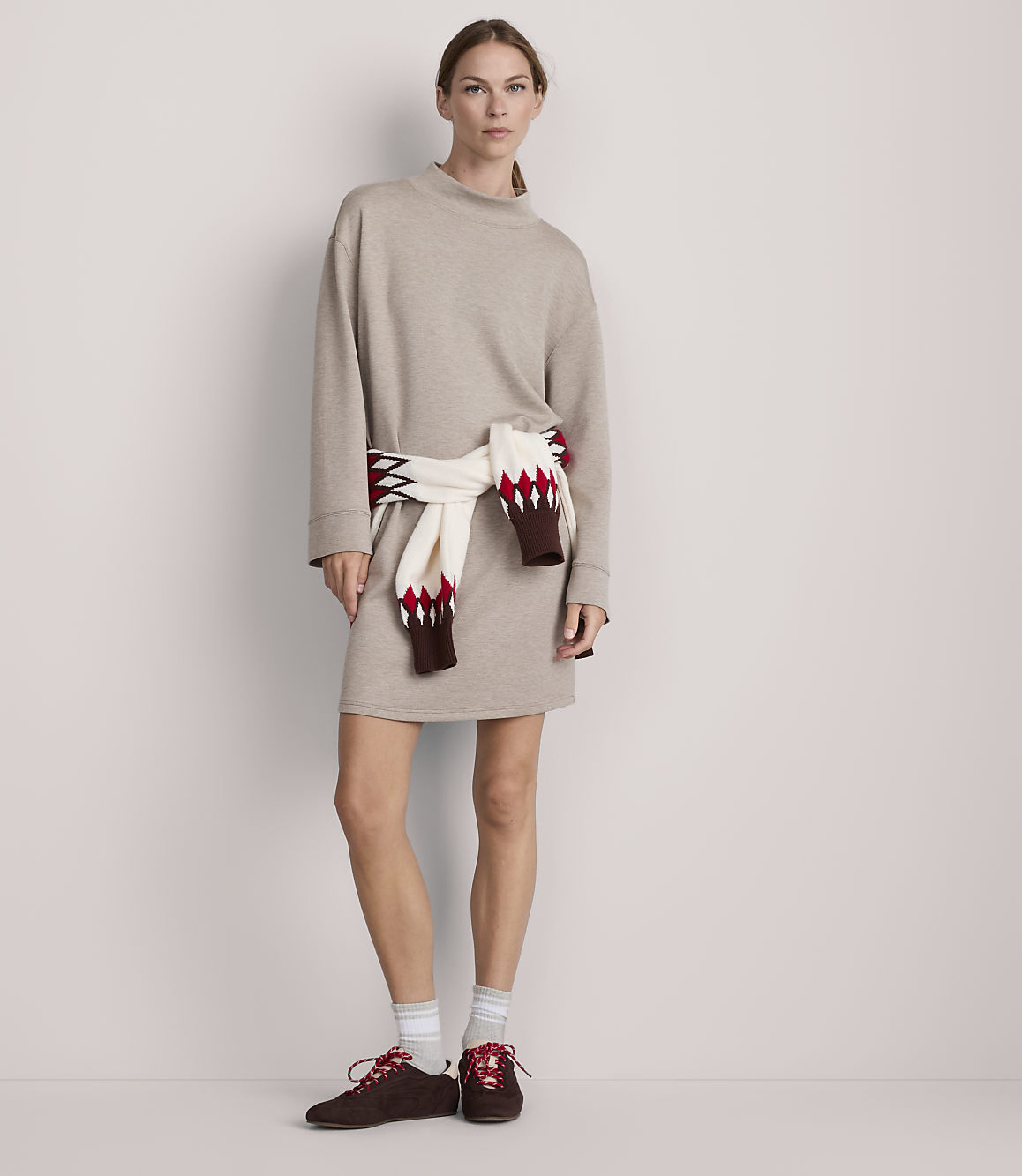 A woman in a beige sweater dress and red sneakers stands against a plain, light-colored wall, with a patterned sweater tied around her waist.