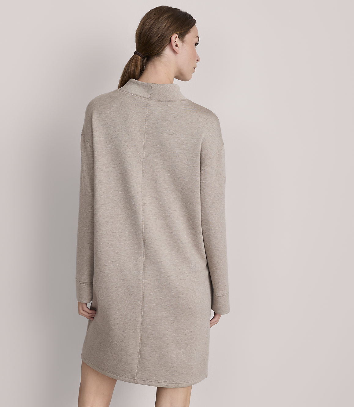 A woman in a beige sweater dress stands against a plain, light gray background.