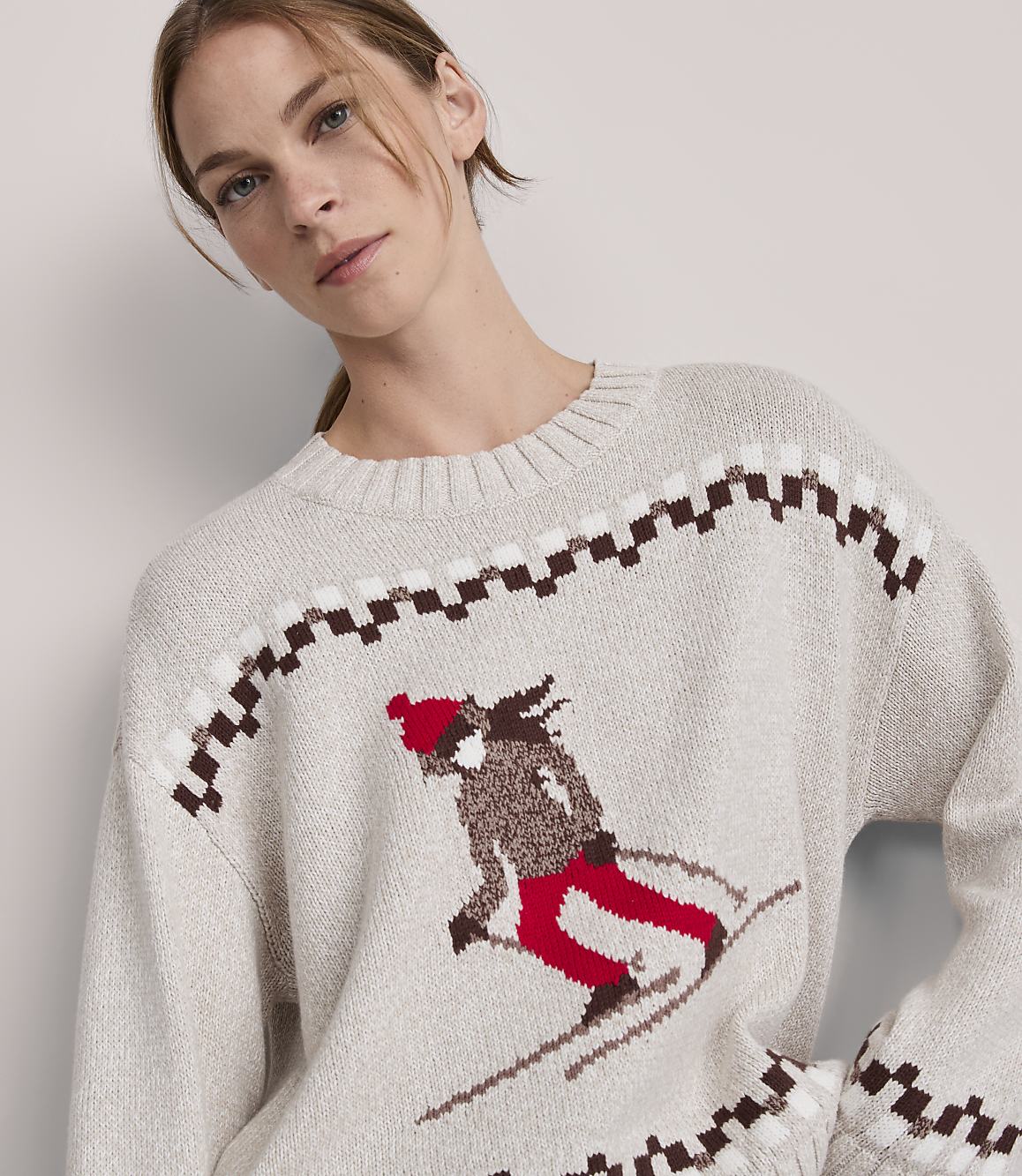 A woman models a cream-colored sweater with a skiing graphic and a checkered pattern, set against a plain, light background.