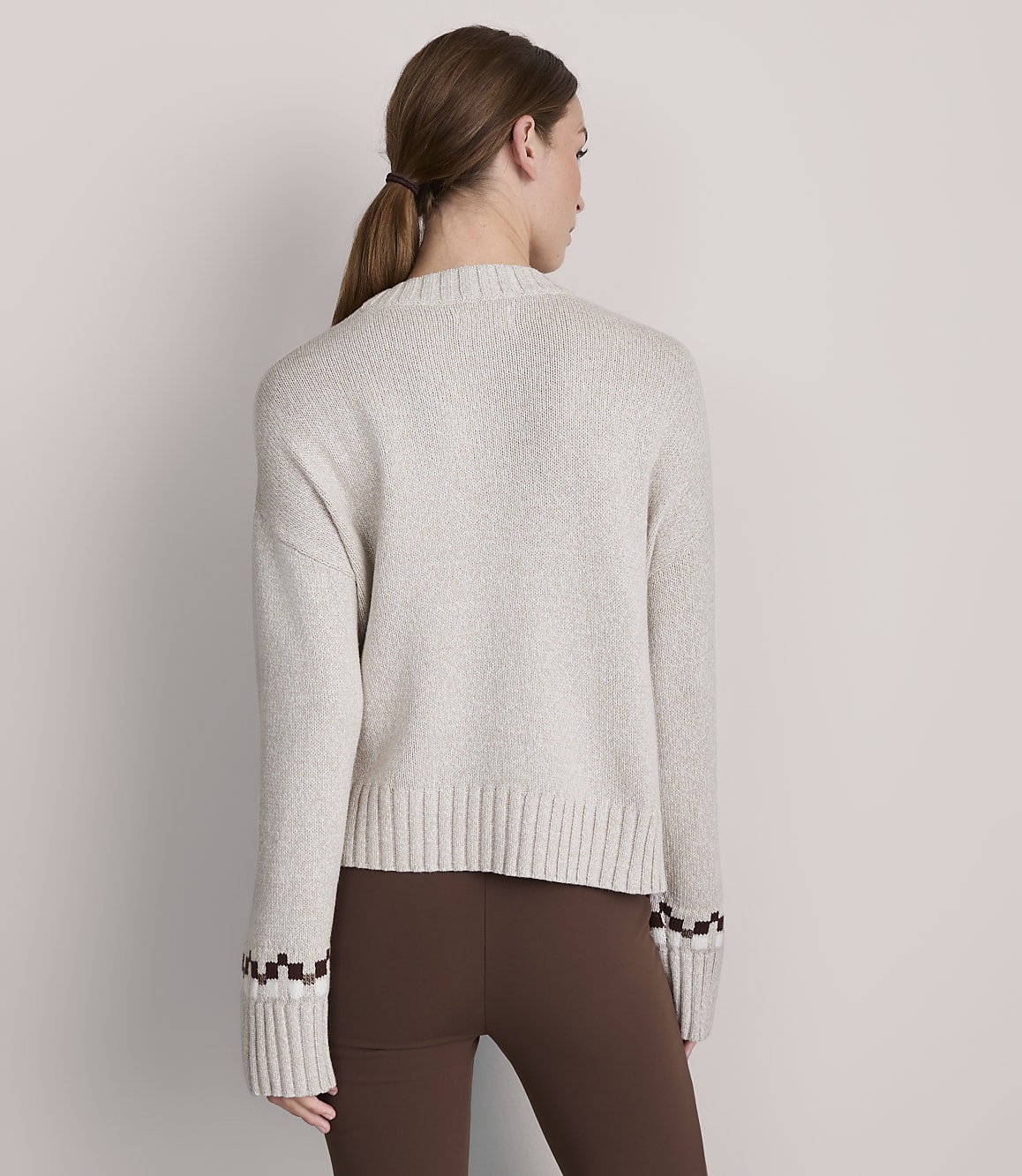 A woman in a light-colored sweater and brown leggings stands against a plain gray background.