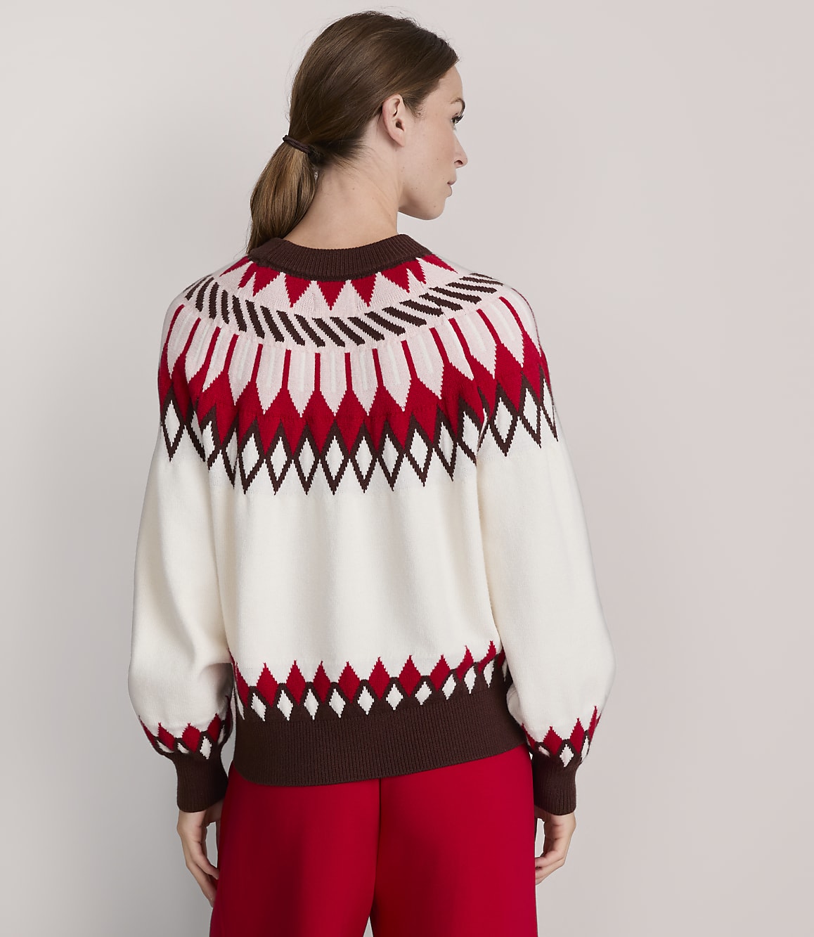 A woman in a cream, red, and brown patterned sweater stands against a plain, light gray background.