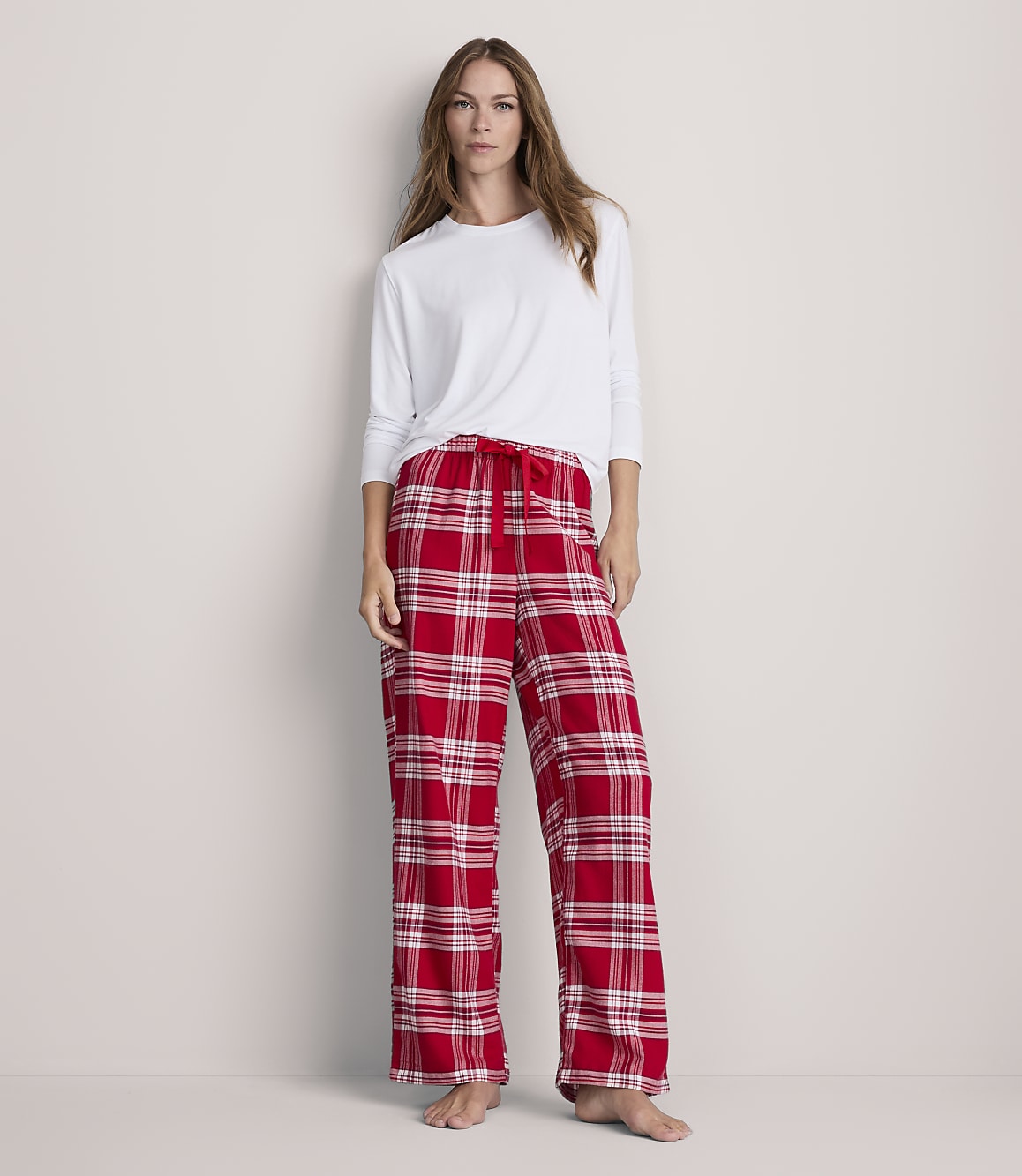 A woman stands in red plaid pajama bottoms and a white long-sleeved shirt against a plain, light-colored wall.