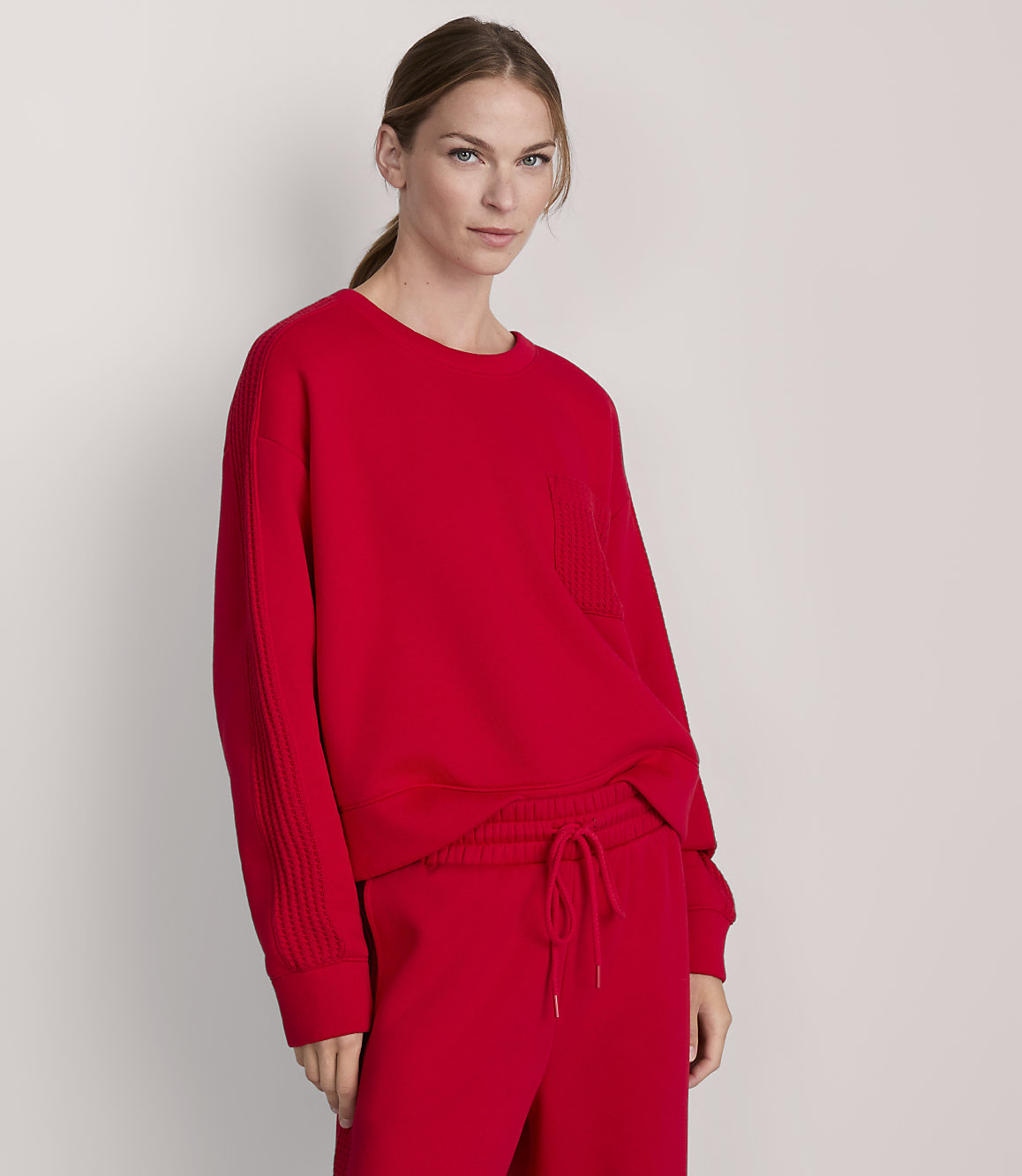 A woman in a red sweatshirt and pants stands against a plain gray background.