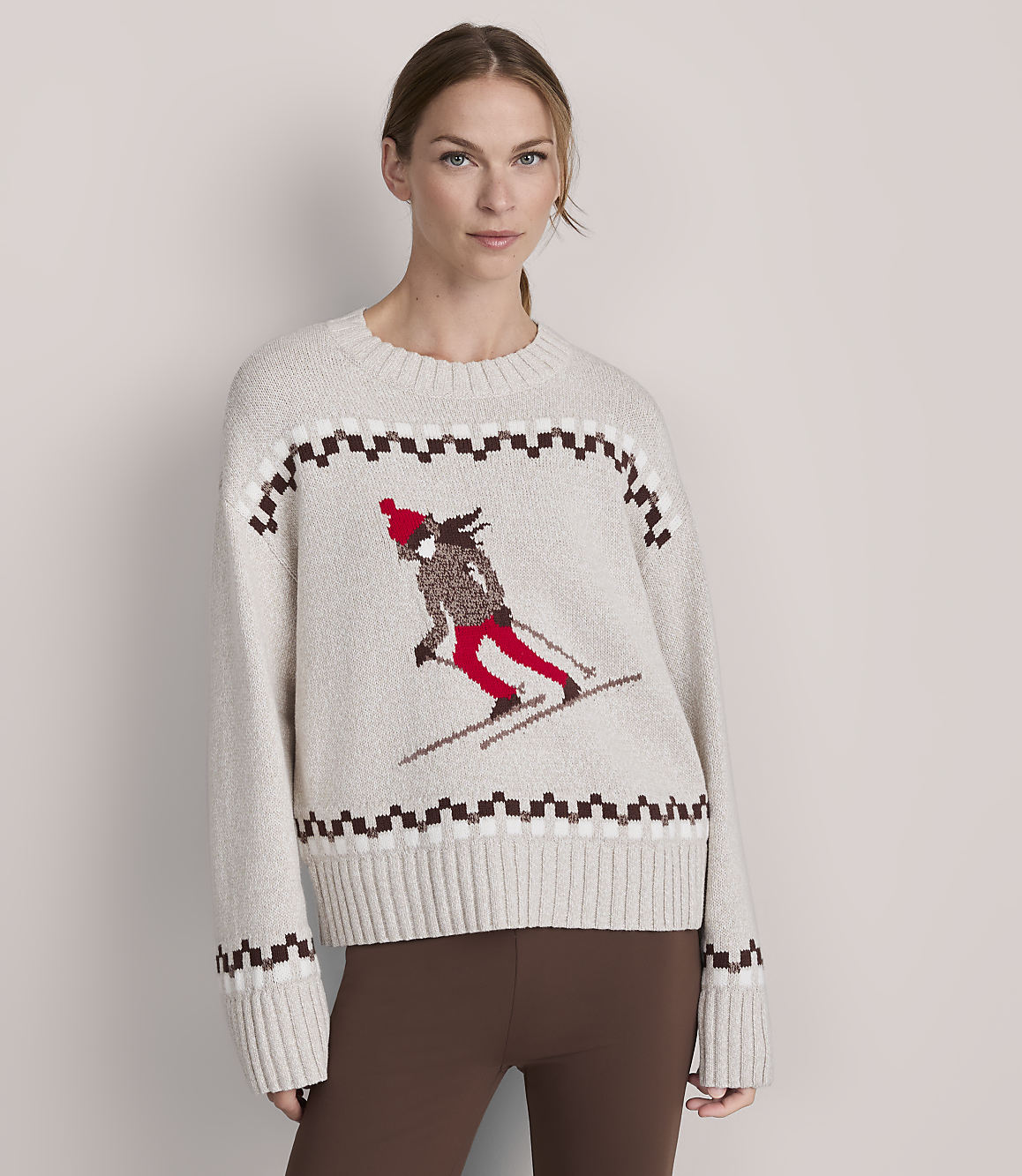 A woman in a cream-colored sweater with a skiing graphic stands against a plain, light gray background.