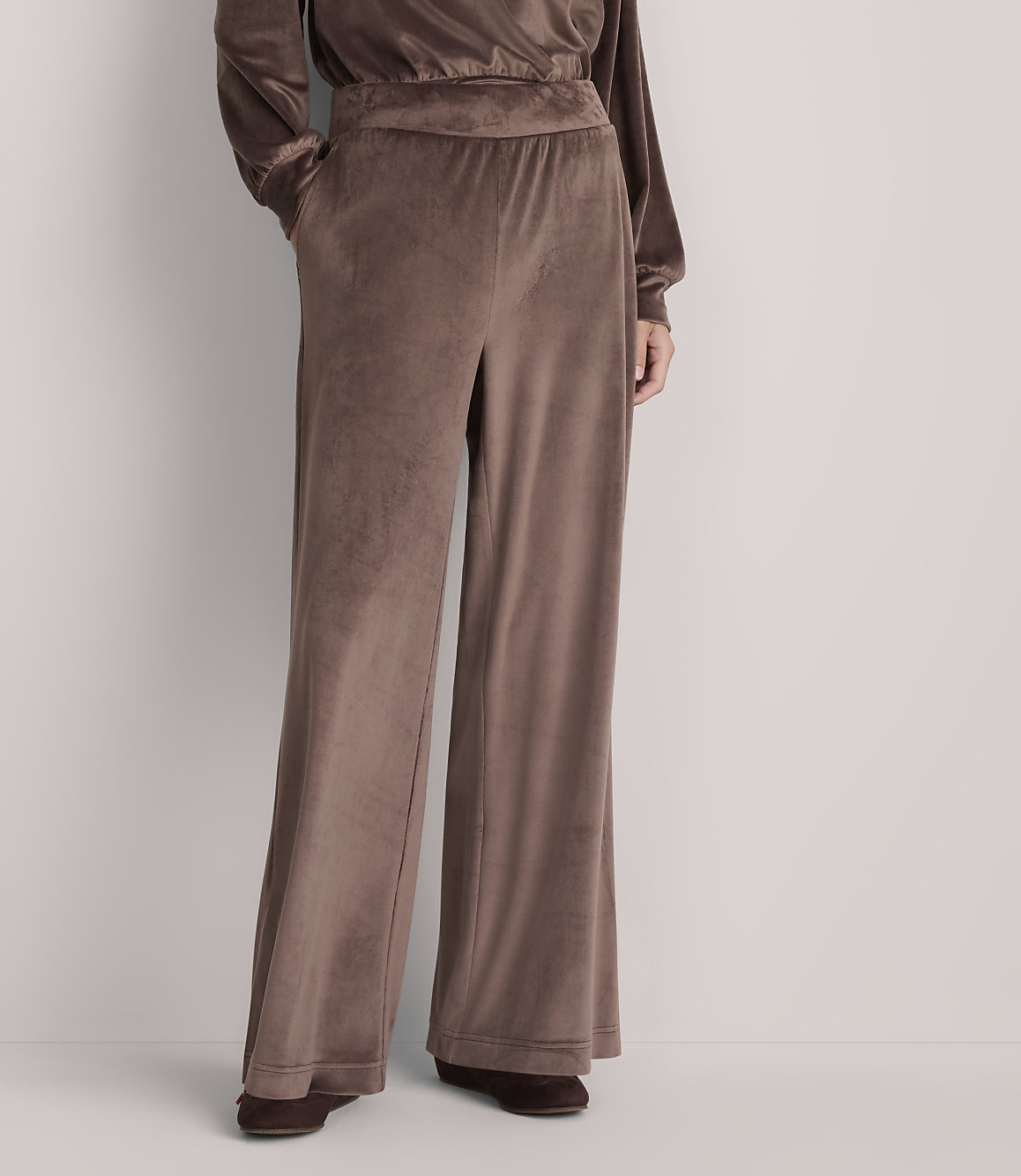 A person stands in loose, brown velvet pants with a plain white background.