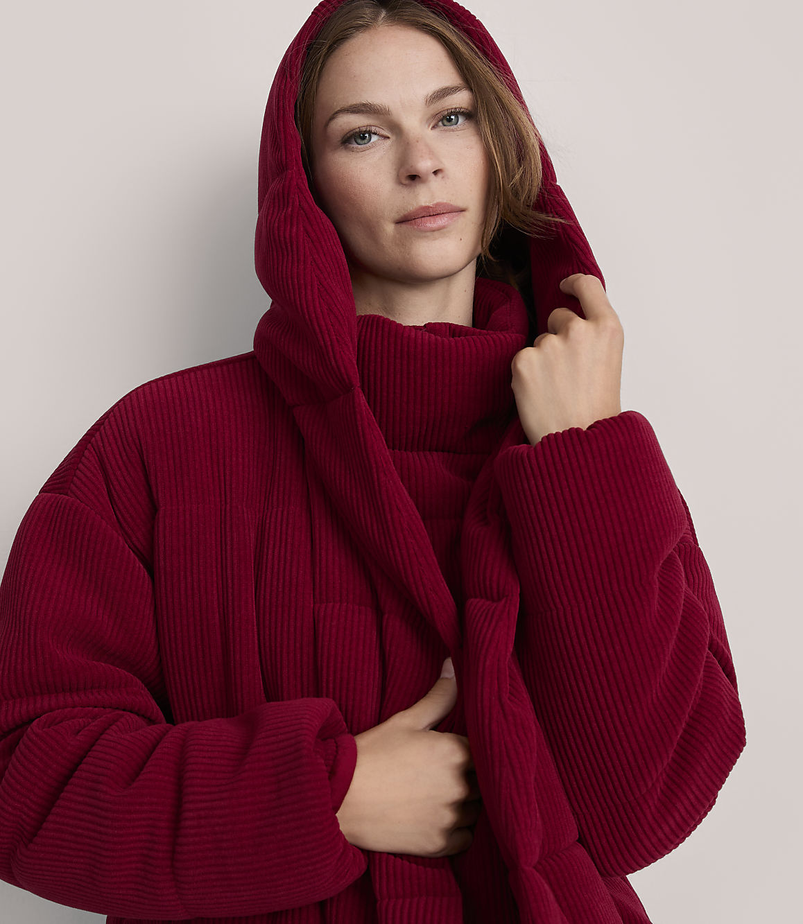 A woman in a red hooded jacket stands against a plain gray background.