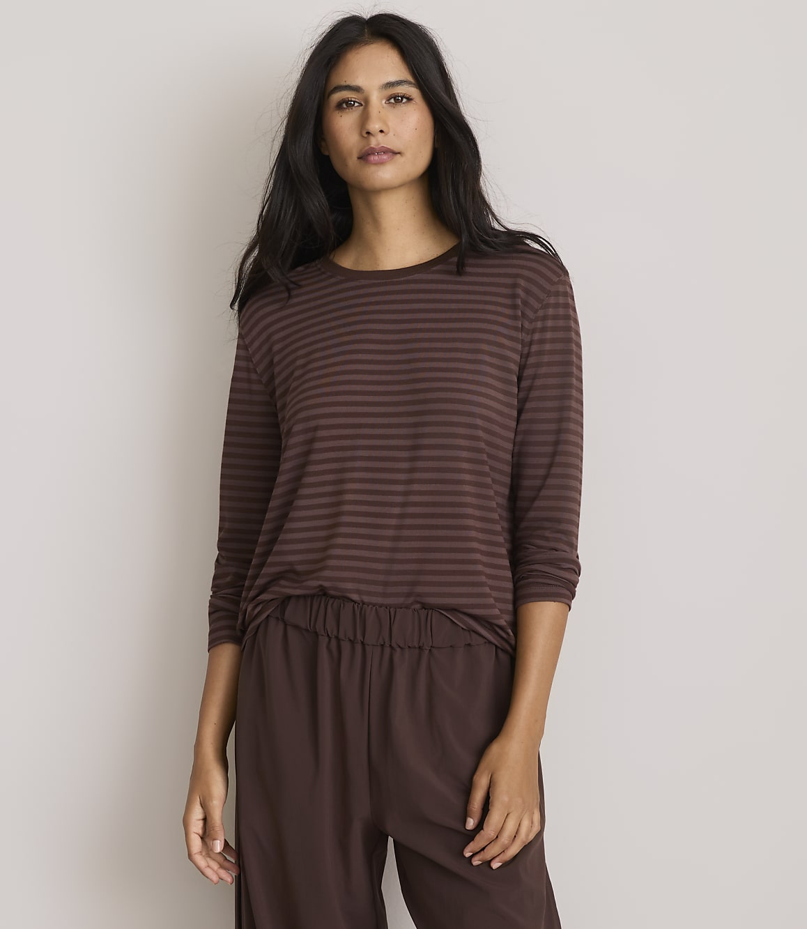 A woman with long dark hair stands in the foreground wearing a brown striped long-sleeved shirt and brown pants, against a plain, light gray background.