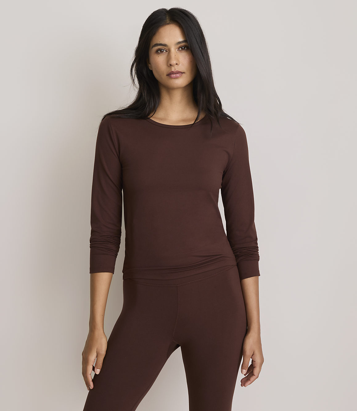 A woman in a brown long-sleeved shirt and leggings stands against a plain, light gray background.