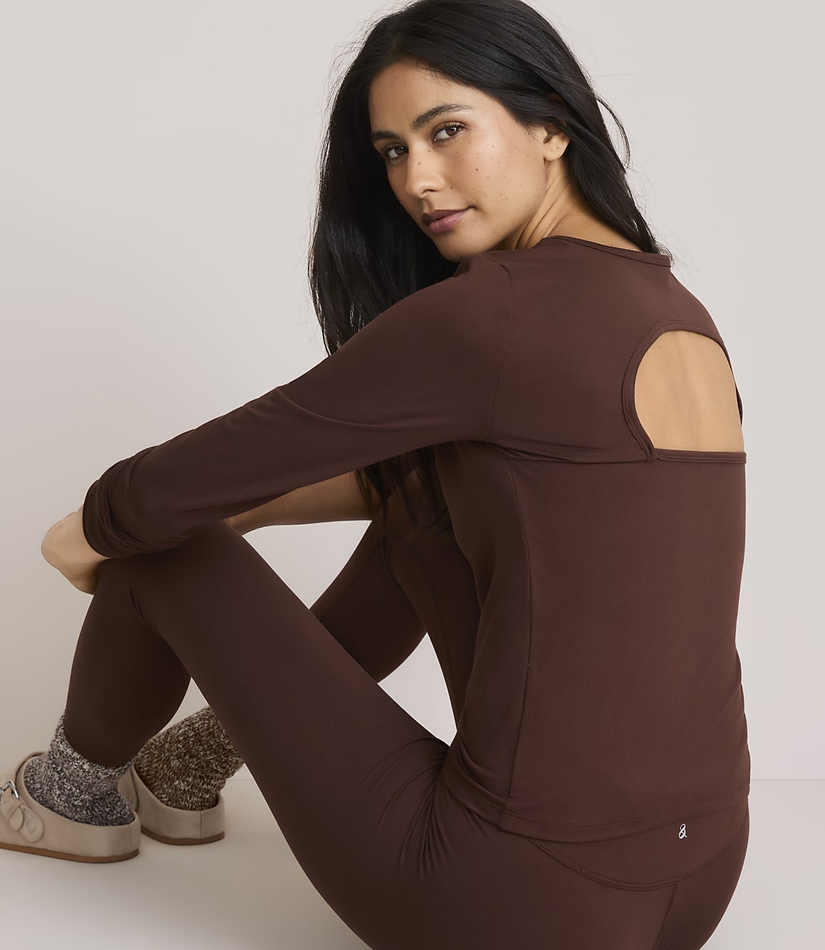 A woman in a brown long-sleeved shirt with an open back and matching leggings sits on a plain background, wearing fuzzy socks and slippers.