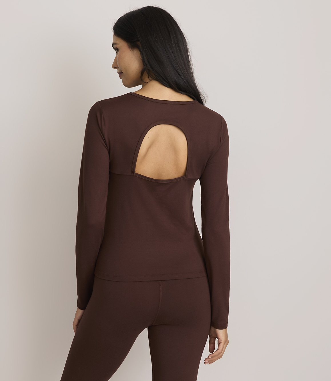 A woman in a brown long-sleeved shirt with a cutout back and matching leggings stands against a plain gray background.