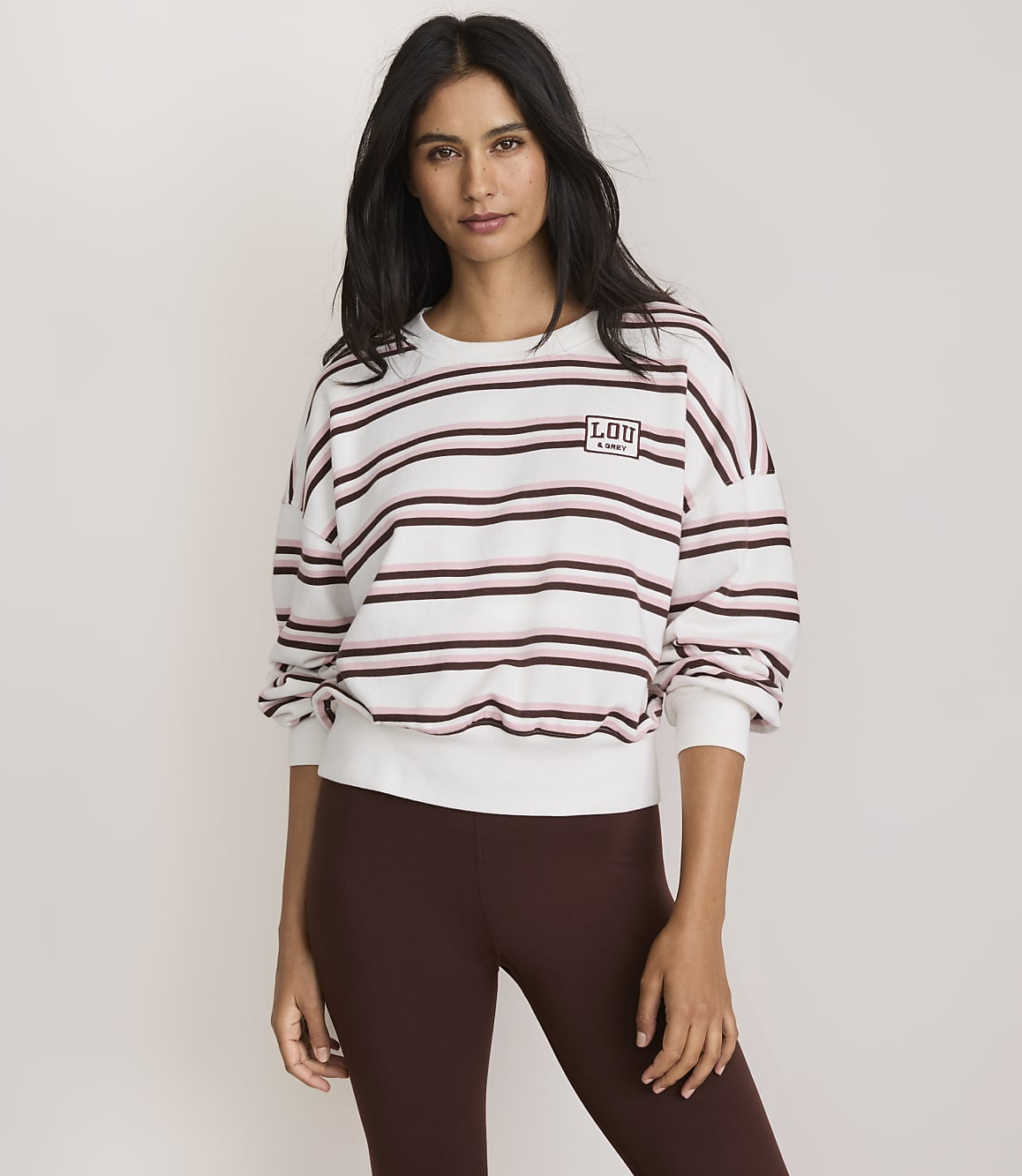 A woman in a striped sweatshirt and brown leggings stands against a plain, light-colored background.