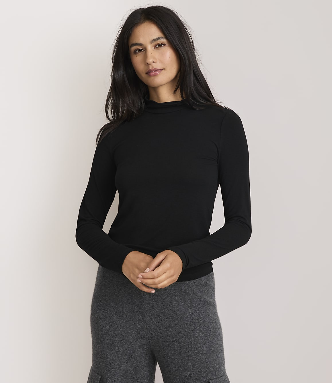 A woman in a black turtleneck and gray pants stands against a plain white background.