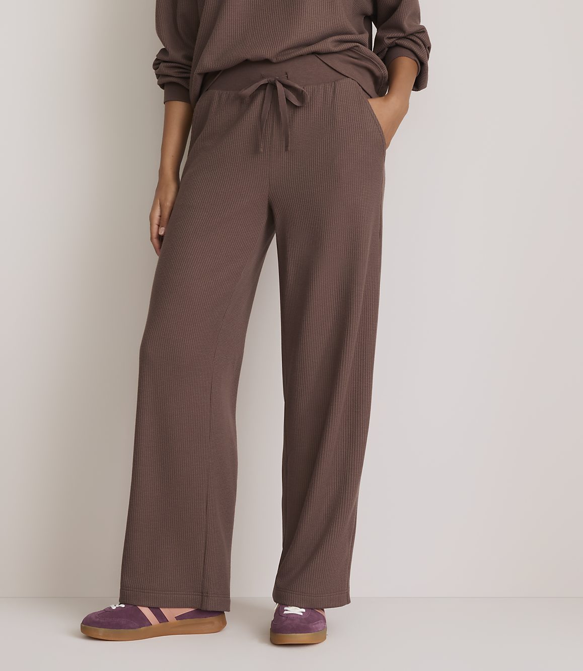 A person stands in brown waffle-knit pants and purple sneakers against a plain, light background.