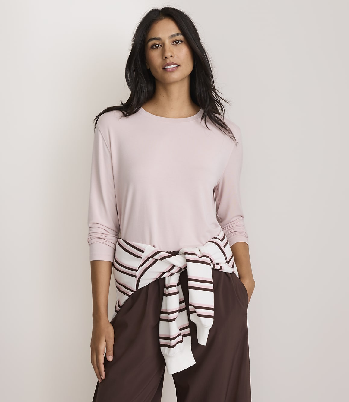 A woman in a pink long-sleeved shirt and brown pants stands against a plain, light-colored background, with a striped sweater tied around her waist.