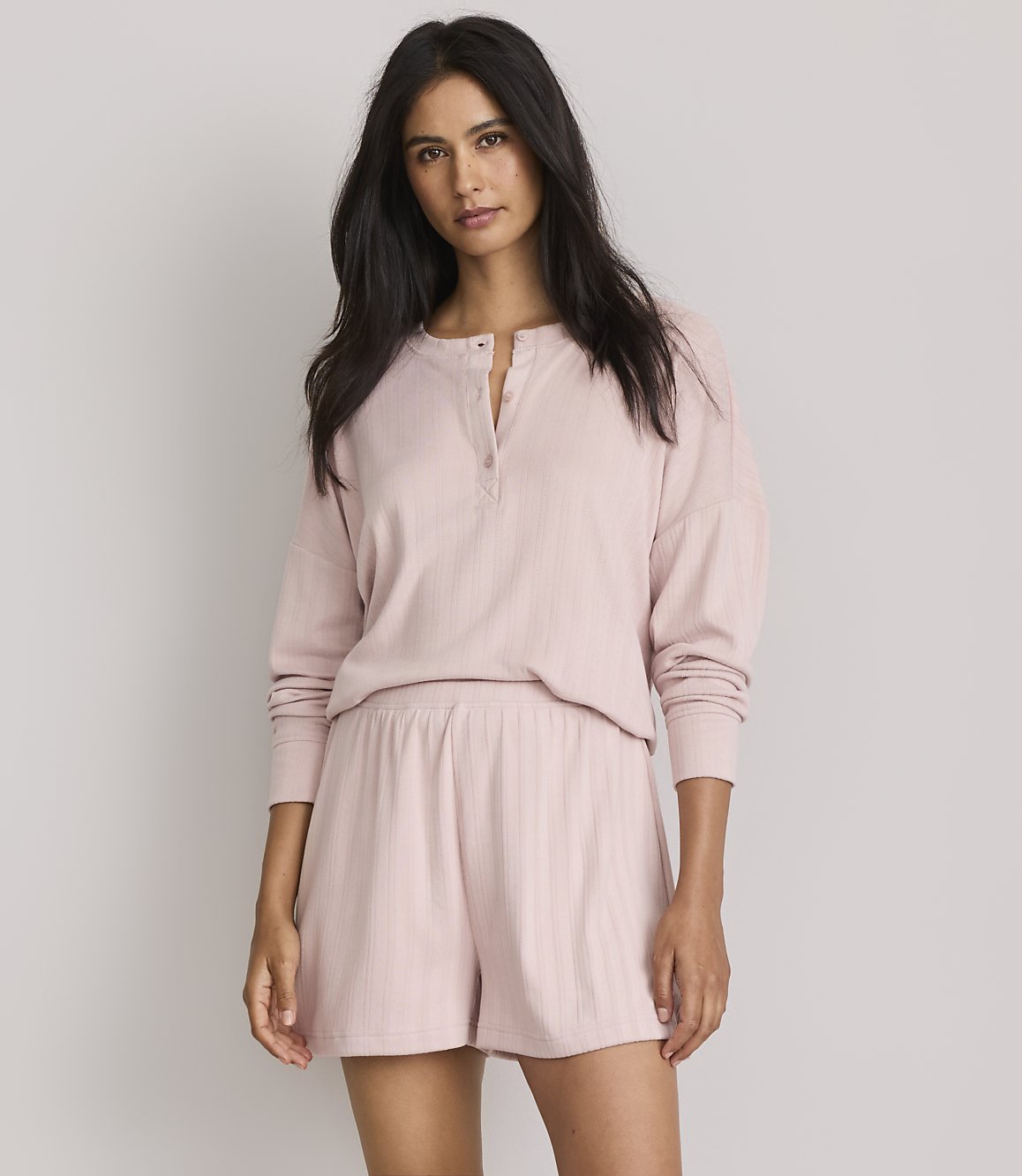 A woman in a pink pajama set stands against a plain gray background.