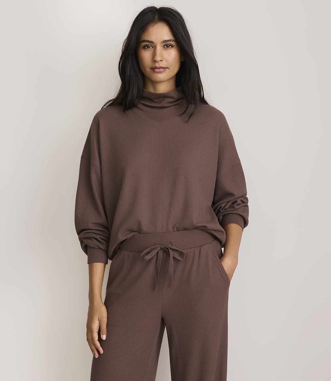 A woman in a brown turtleneck sweater and matching pants stands against a plain, light-colored background.