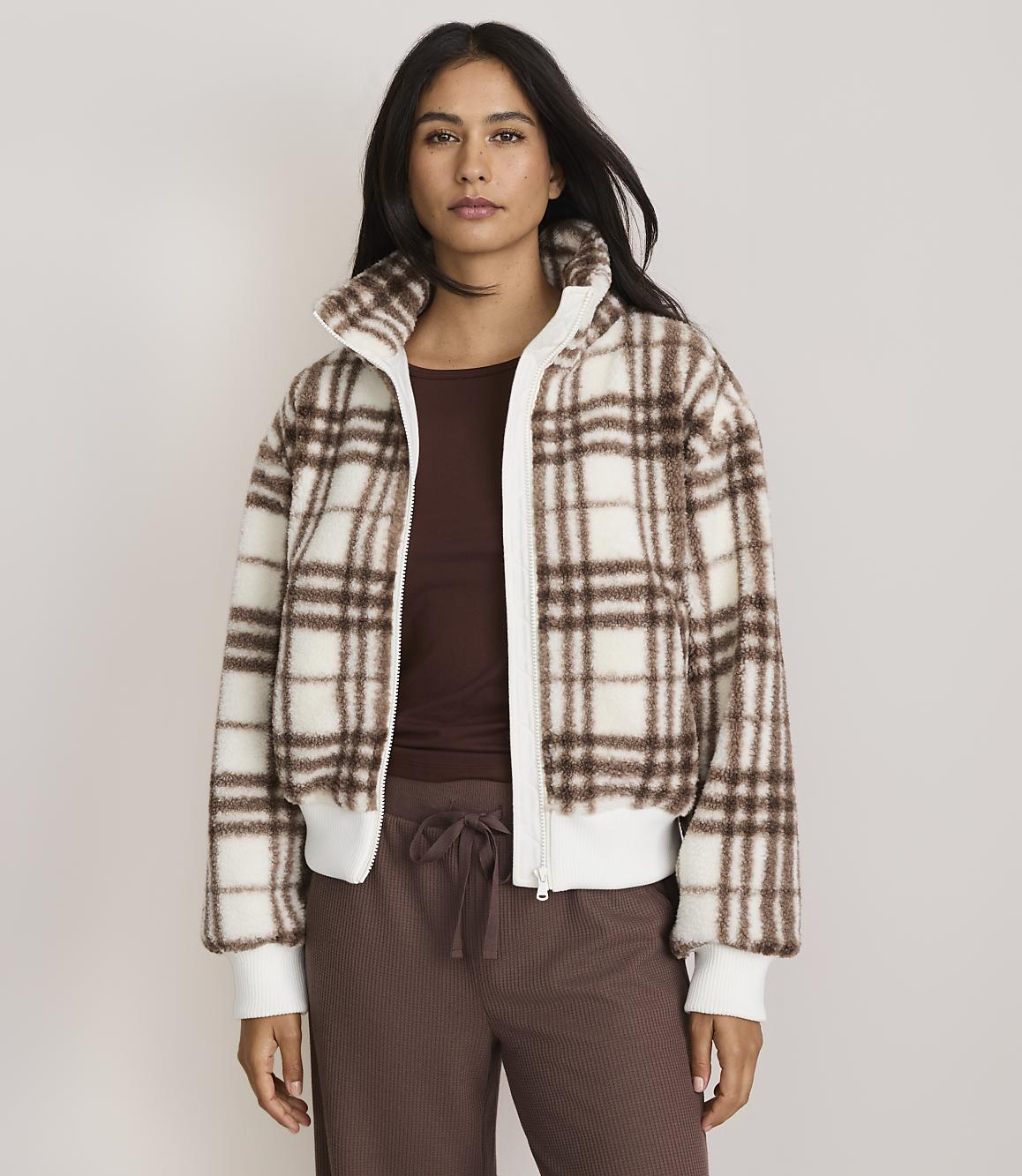 A woman in a plaid jacket and brown pants stands against a plain, light-colored background.