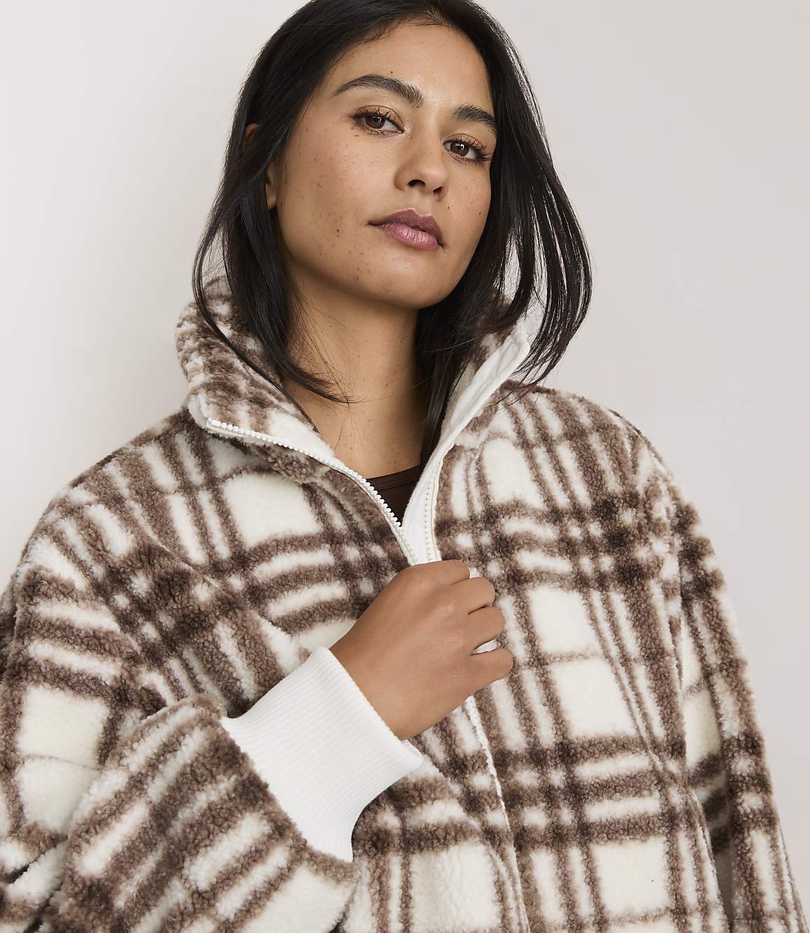 A woman in a brown and white plaid jacket stands against a plain white background.