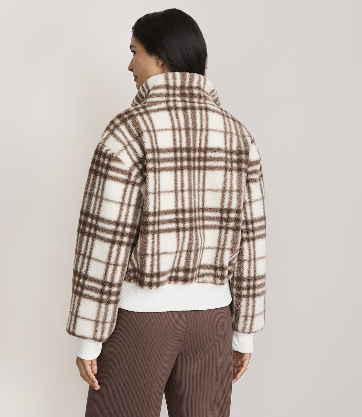 A woman in a brown and white plaid jacket stands against a plain, light-colored background.