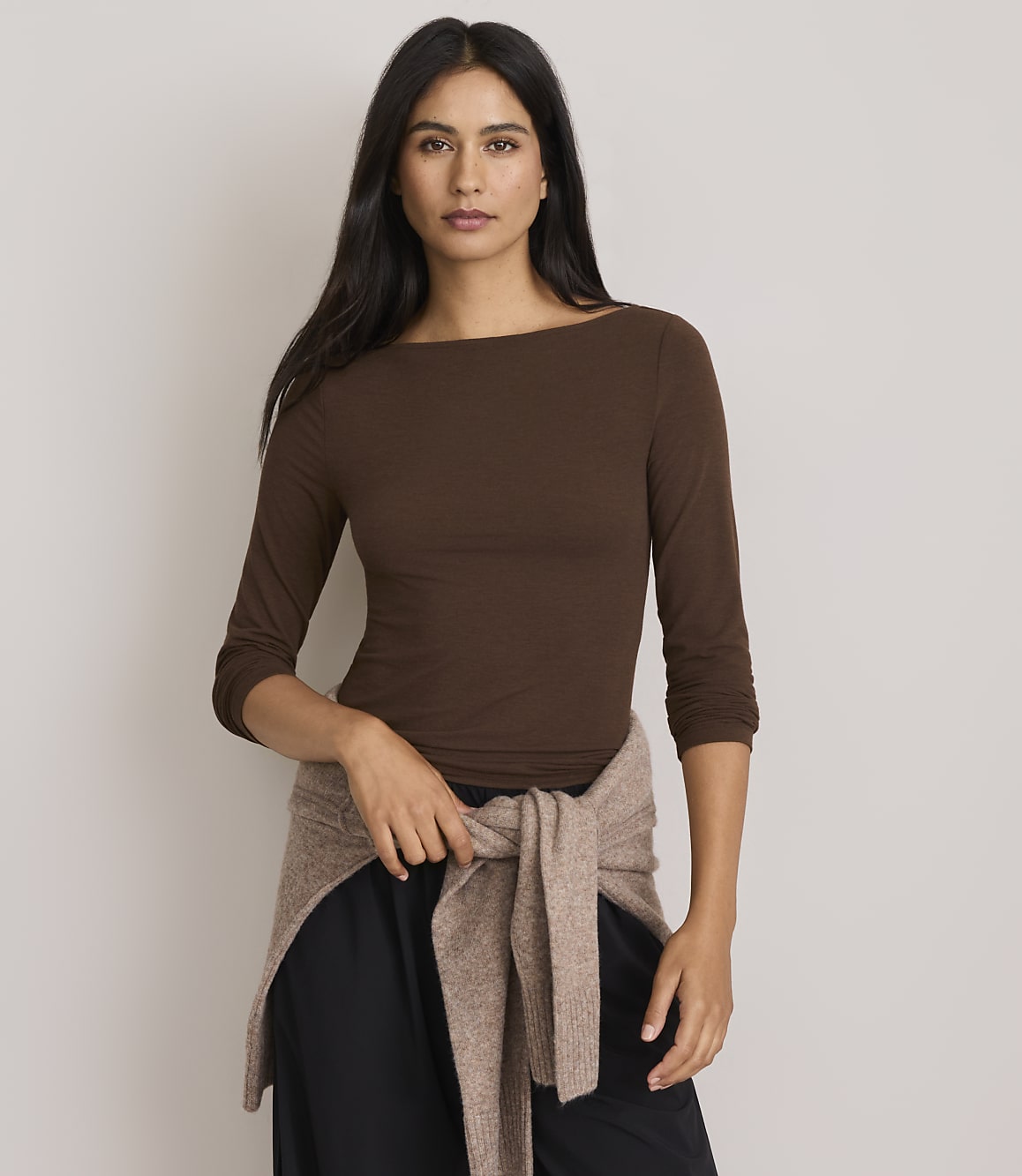 A woman in a brown boatneck shirt and black pants stands against a plain gray background, with a tan sweater tied around her waist.