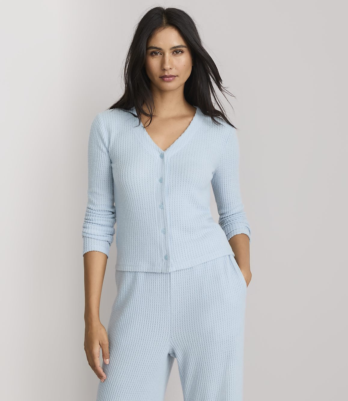 A woman in a light blue ribbed cardigan and matching pants stands against a plain white background.