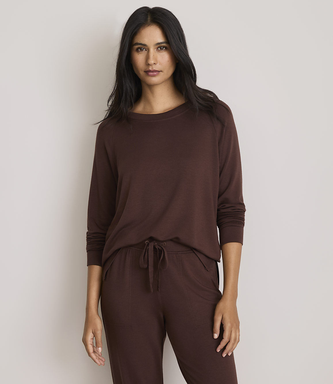 A woman in a brown long-sleeved shirt and pants stands against a plain, light gray background.