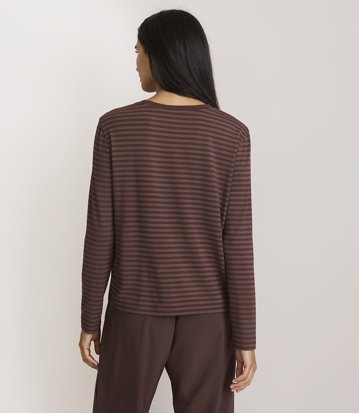 A woman in a brown striped shirt and brown pants stands against a plain, light background.