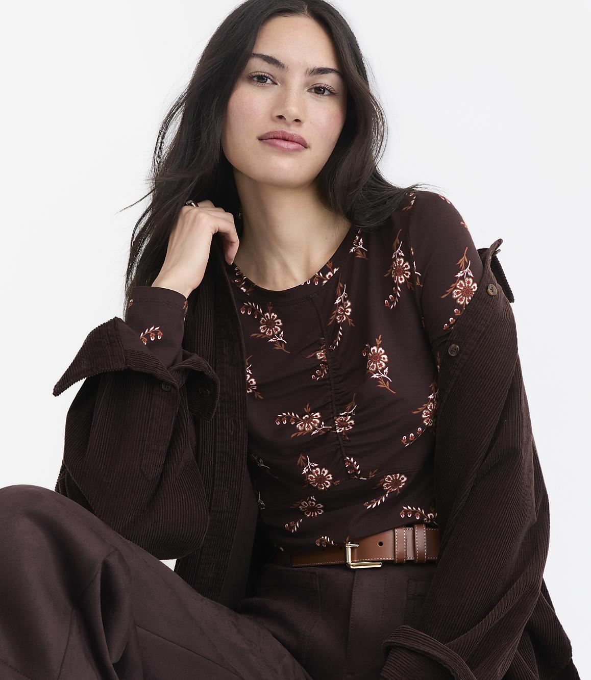 A woman with dark hair and a floral top is seated in front of a plain white background.