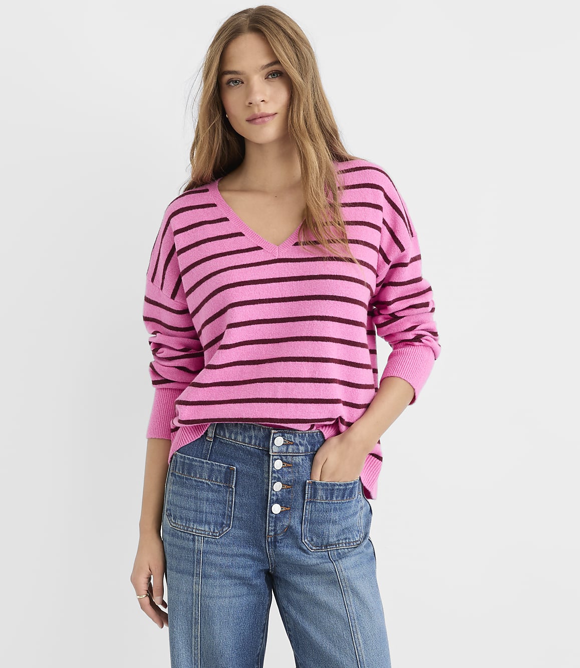 A young woman in a pink and purple striped sweater and blue jeans stands against a plain white background.