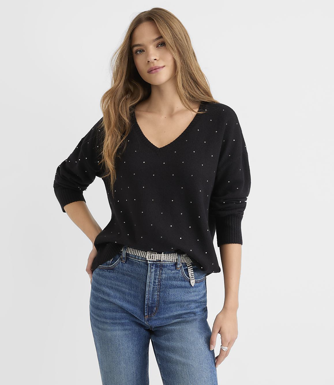 A young woman in a sparkling black v-neck sweater and blue jeans stands against a plain white background.
