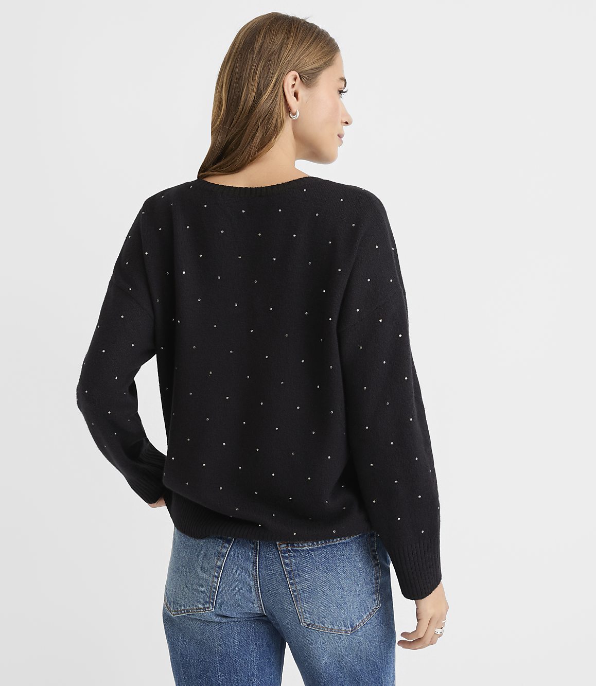 A woman in a black studded sweater and blue jeans stands against a plain white background.