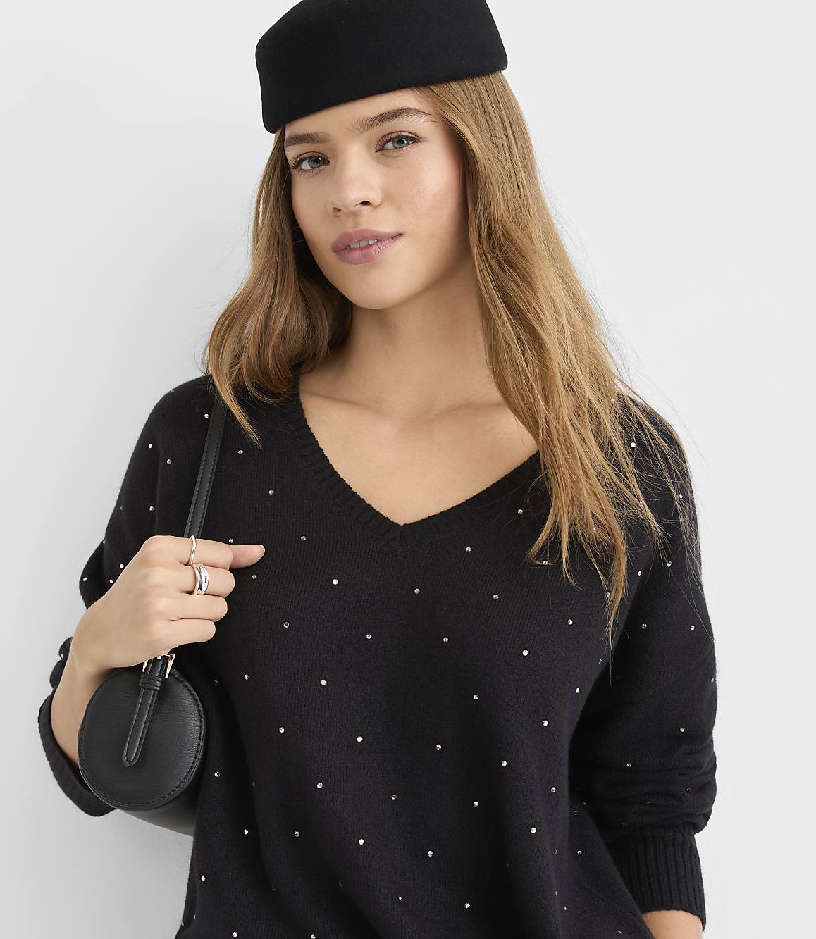 A woman in a black, studded V-neck sweater and a black pillbox hat poses against a plain white background, holding a small black purse.