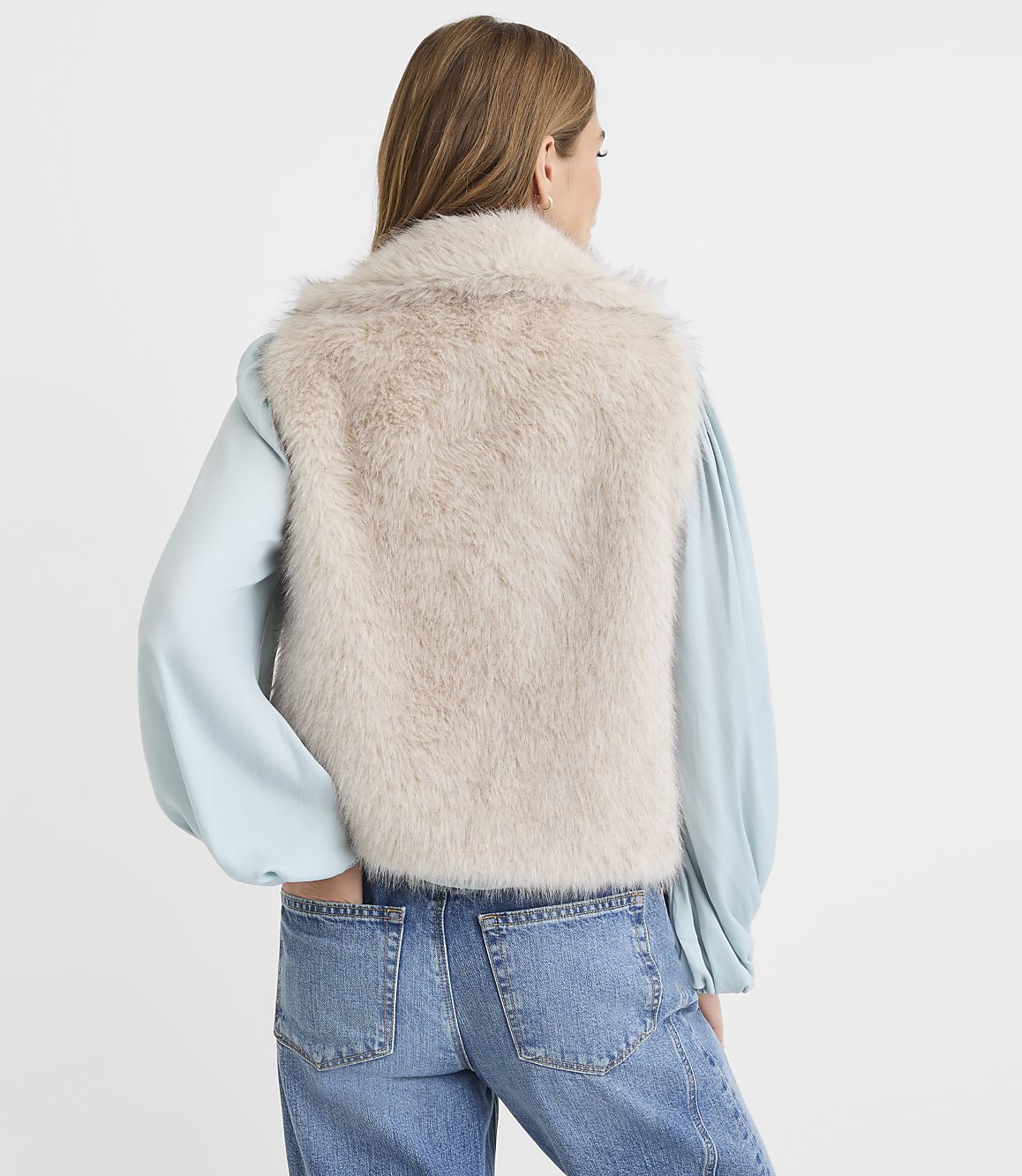 A woman in a light blue blouse and jeans stands with her back to the camera, wearing a fluffy cream-colored vest against a plain white background.
