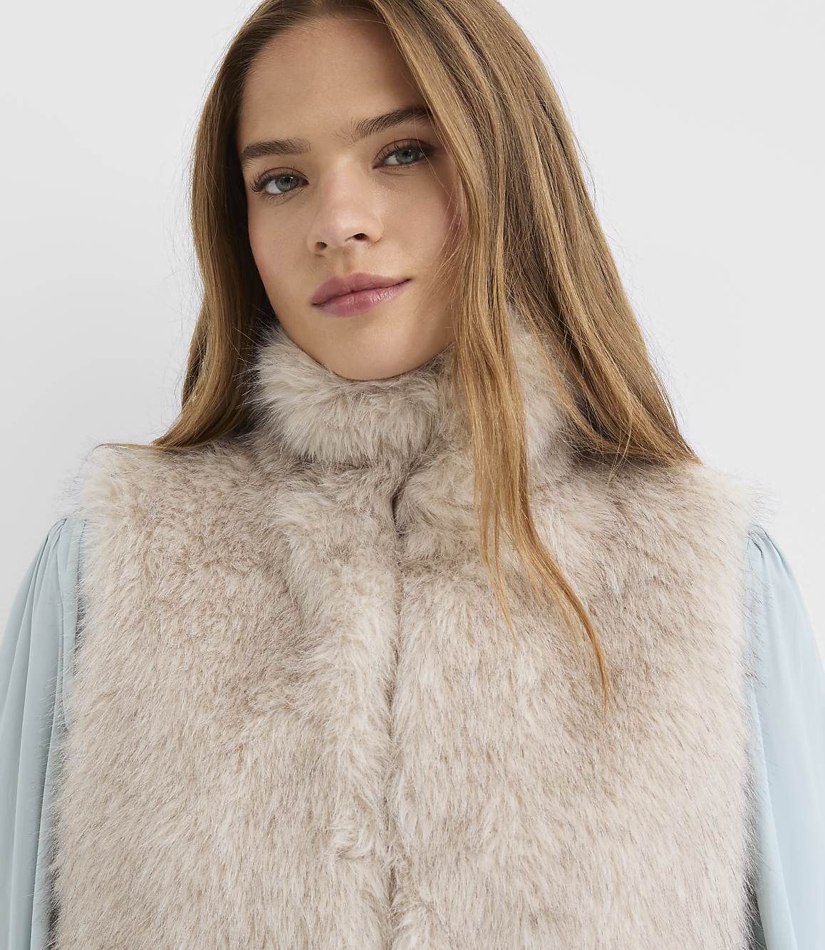 A woman with long blonde hair wears a light beige fur vest over a light blue blouse against a plain white background.
