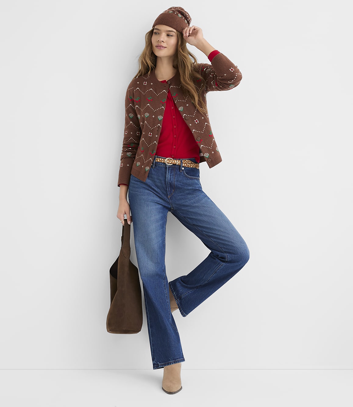 A woman in a brown patterned cardigan, red top, and flared jeans poses against a plain white background, holding a brown tote bag.