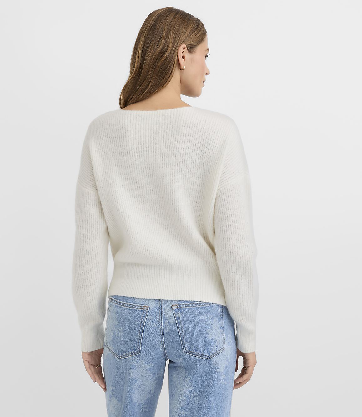 A woman in a white ribbed sweater and light blue jeans with a subtle floral pattern stands against a plain white background.