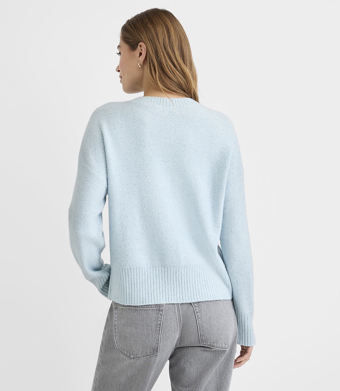 A woman in a light blue sweater and grey jeans stands against a plain white background.
