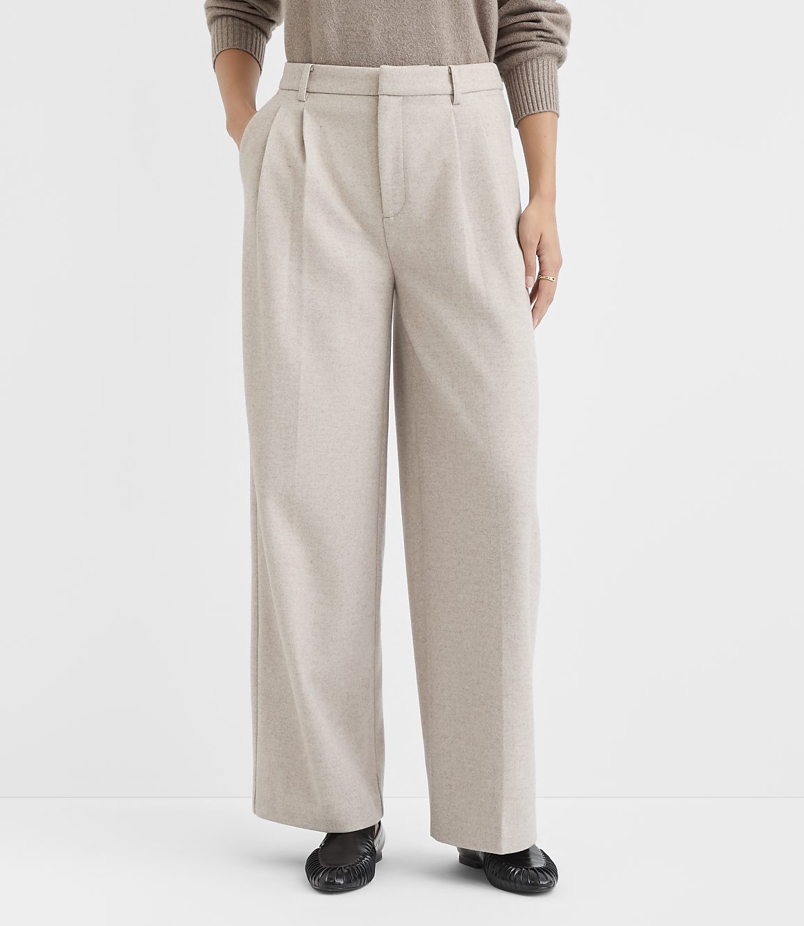A person stands in the foreground wearing beige pleated wide-leg pants and a brown sweater, against a plain white background.