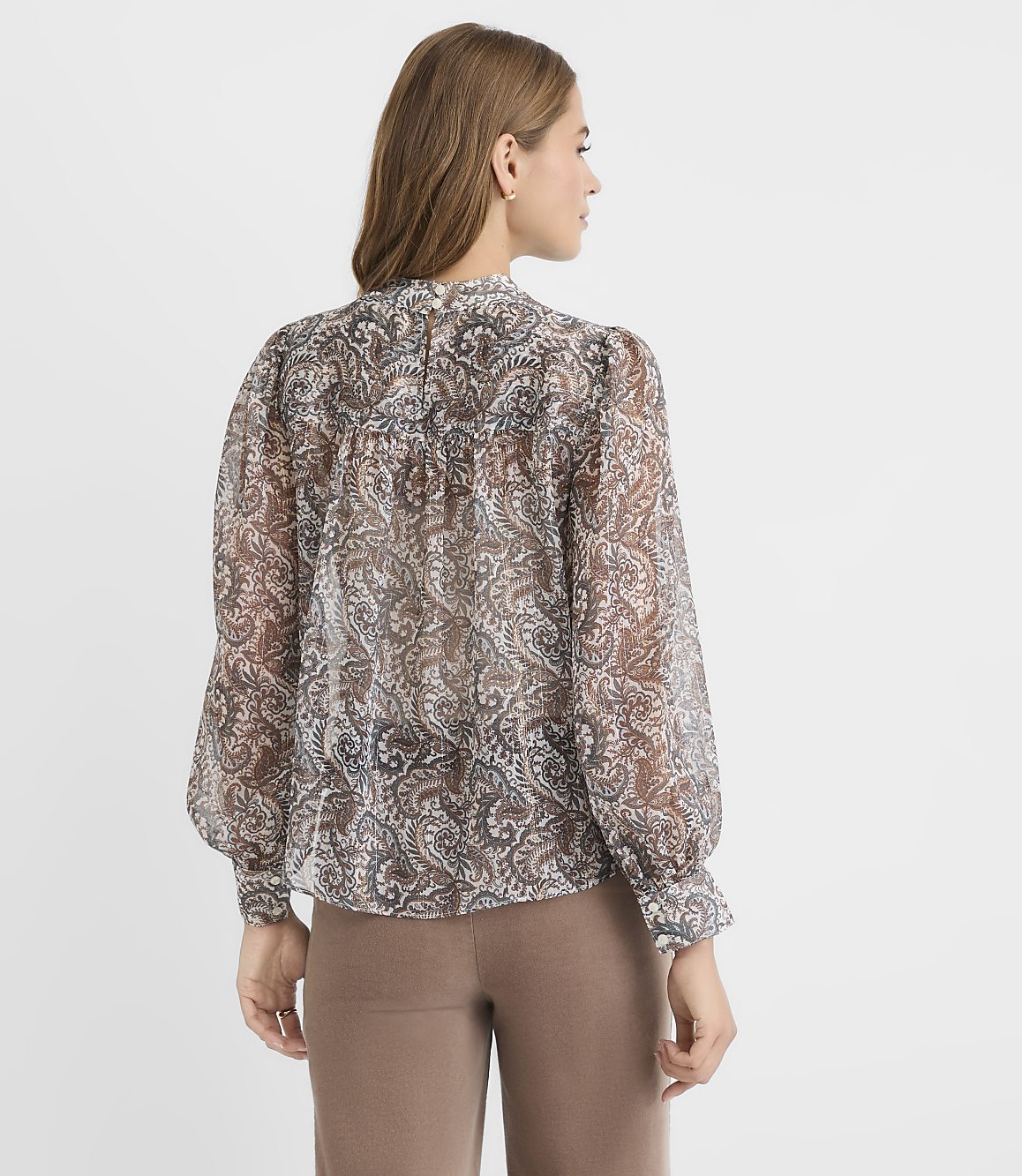 A woman in a patterned sheer blouse and brown pants stands against a plain white background.