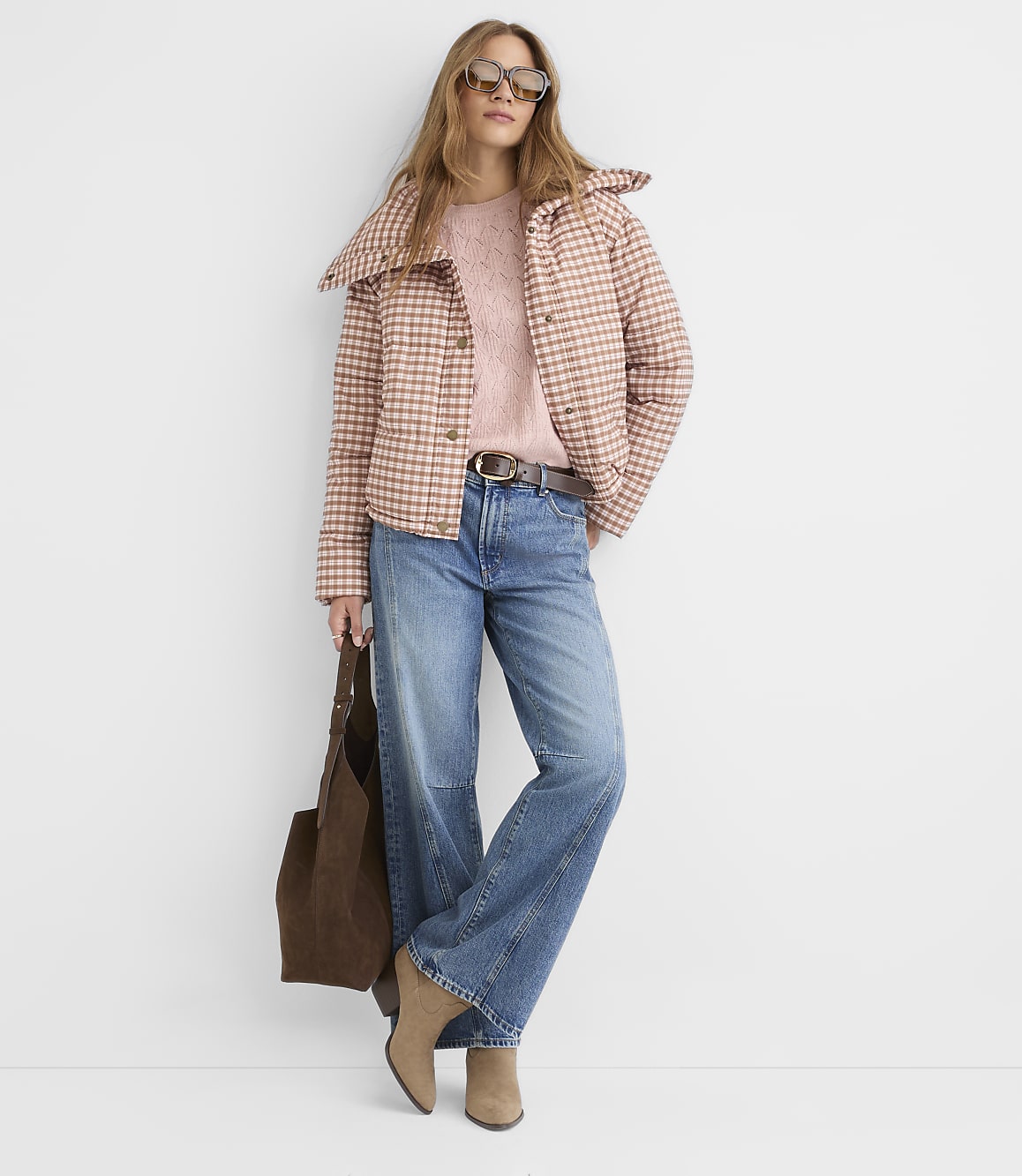 A woman in a pink plaid jacket and jeans stands against a plain white background, holding a brown tote bag.