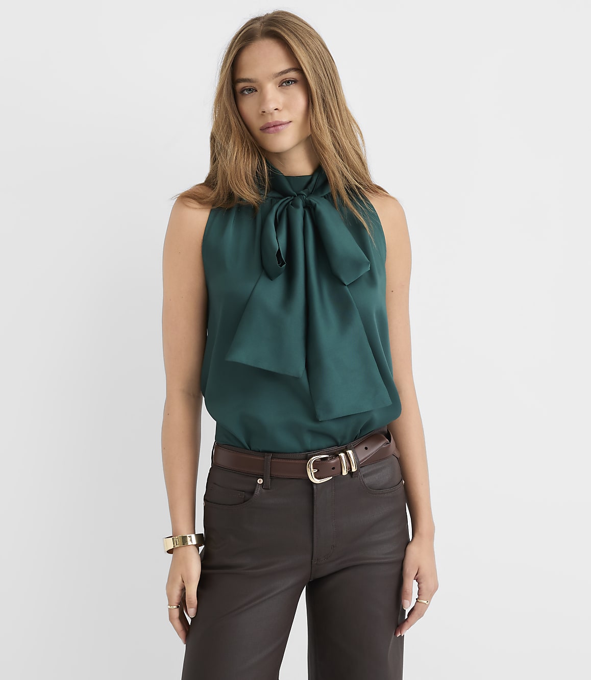 A woman in a teal bow-neck blouse and dark pants stands against a plain white background.