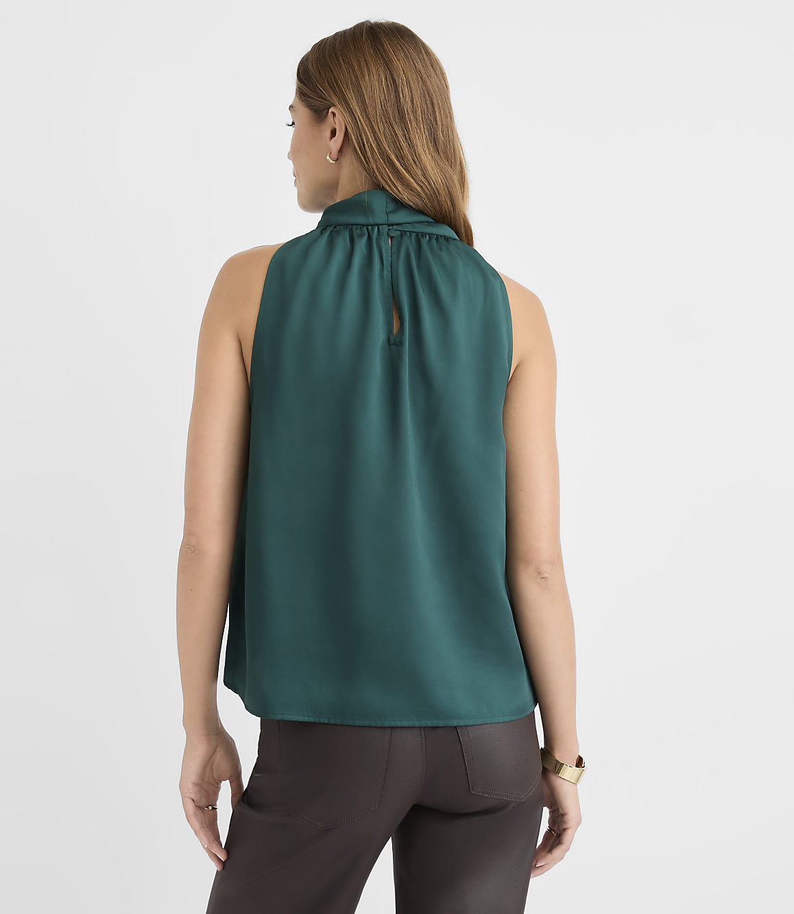 A woman in a teal sleeveless top and brown pants stands against a plain white background.