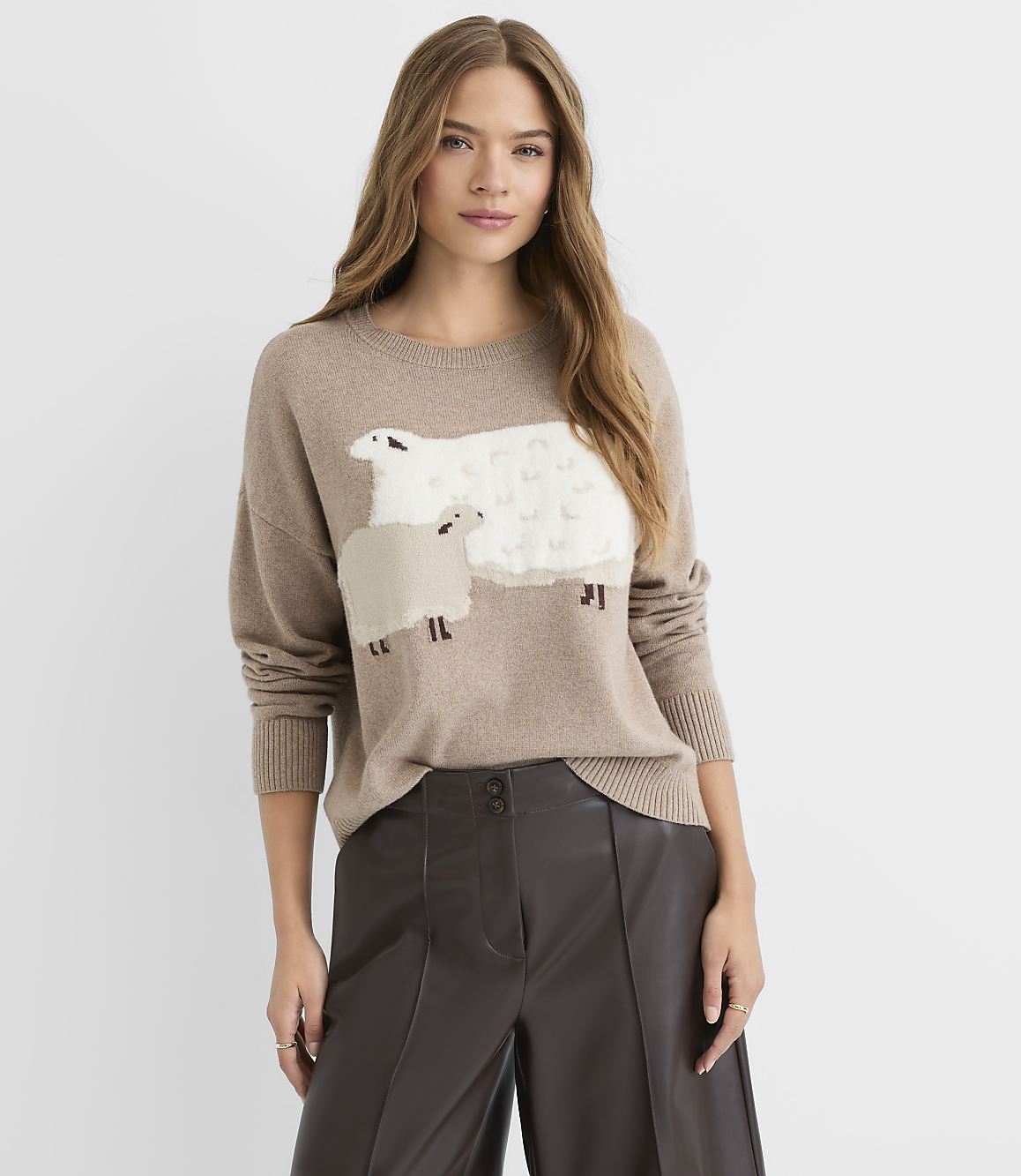 A woman in a tan sweater with sheep appliqu&eacute;s stands against a plain, light background.