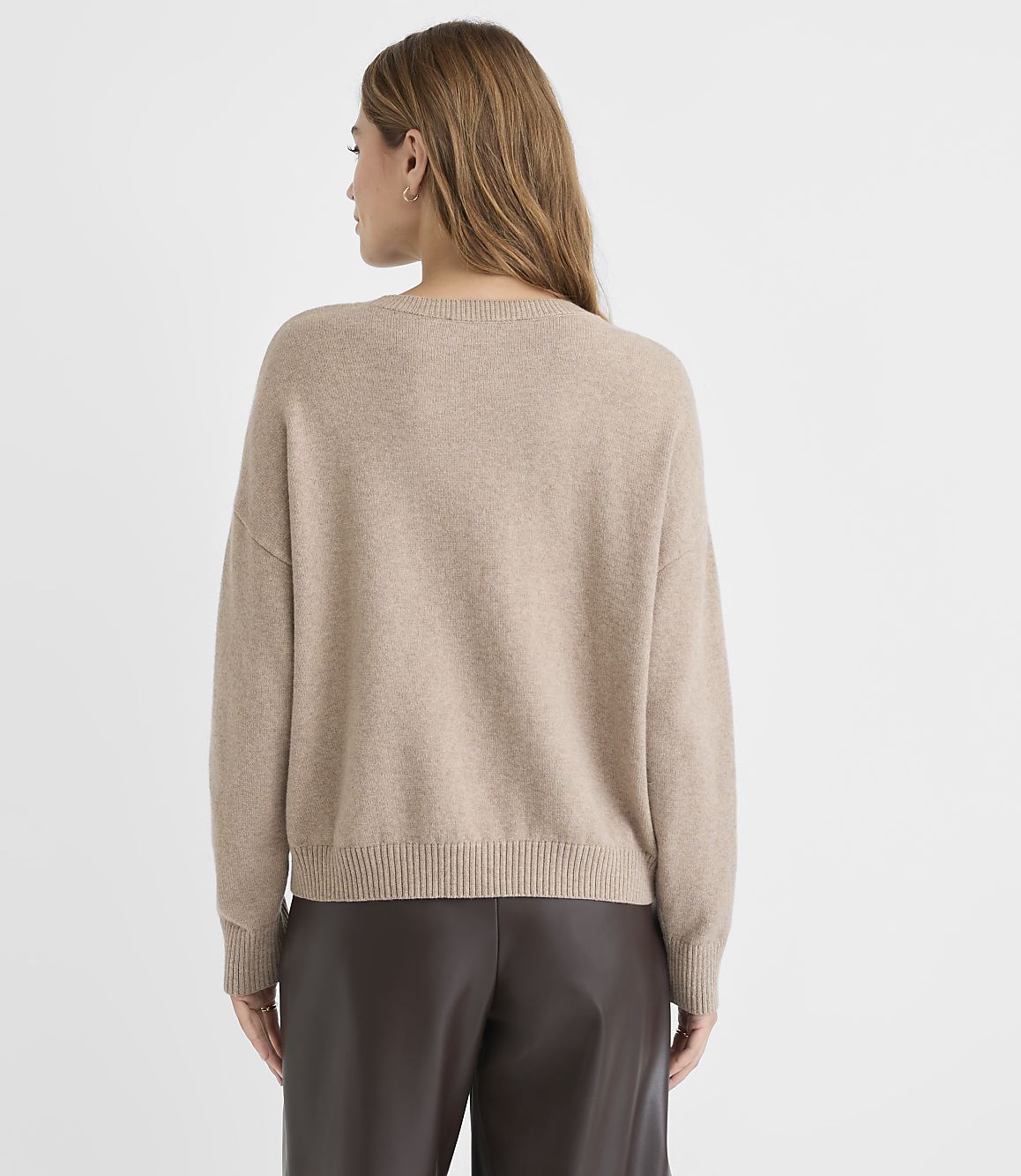 A woman in a beige sweater and brown leather pants stands against a plain white background.