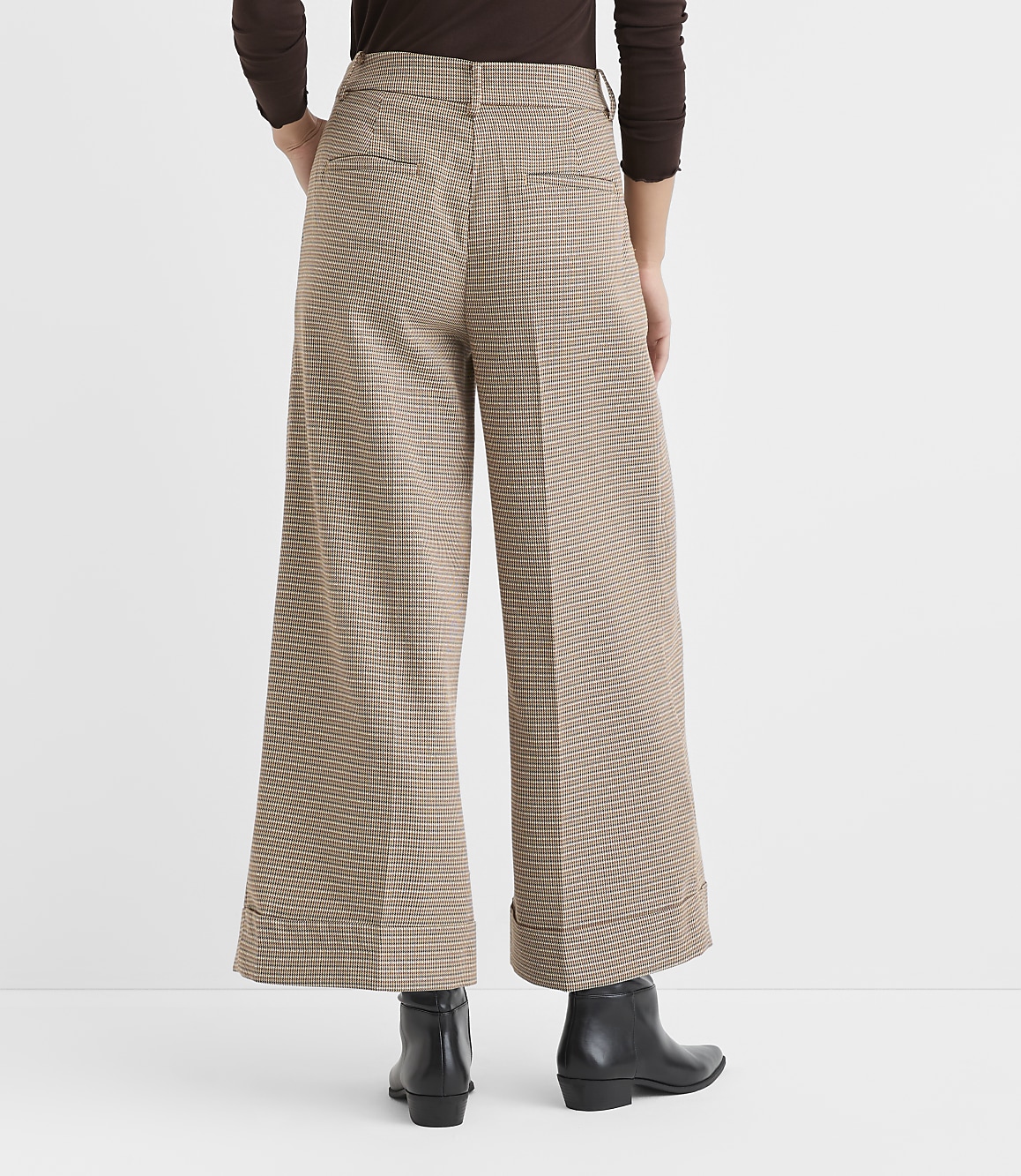A person stands in the foreground wearing wide-leg, checkered pants and black boots, with a plain brown background.