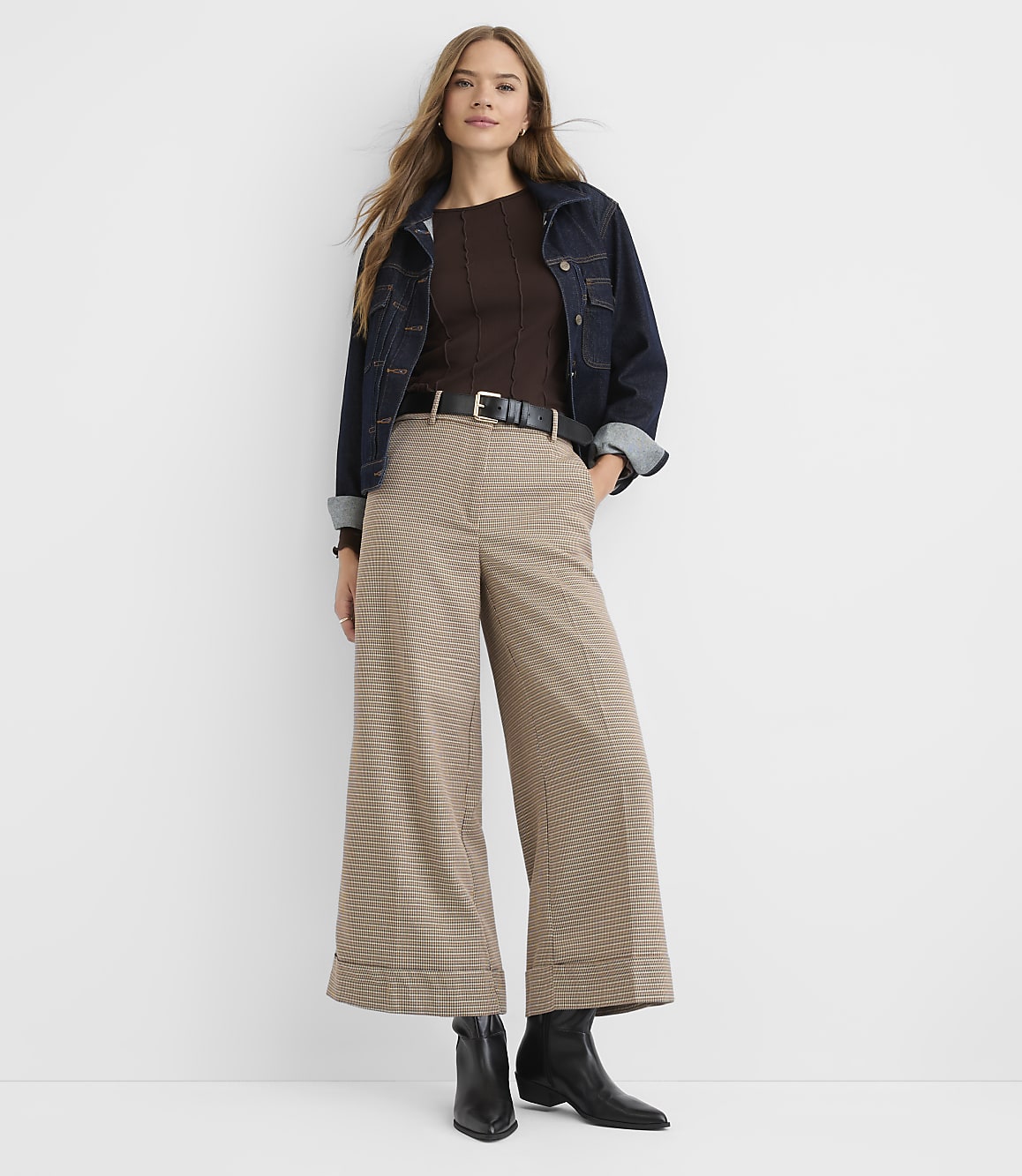 A woman in a brown ribbed top, dark denim jacket, and wide-leg plaid pants stands against a plain white background.