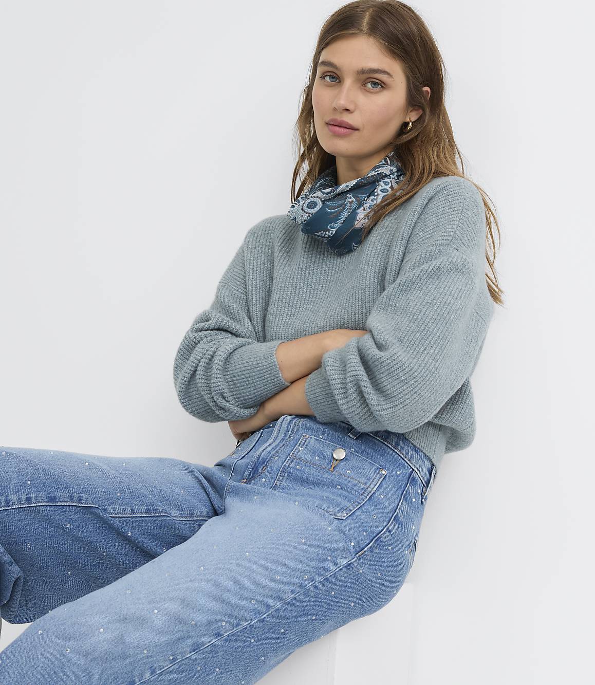 A woman in a light blue sweater and embellished jeans poses against a plain white background.
