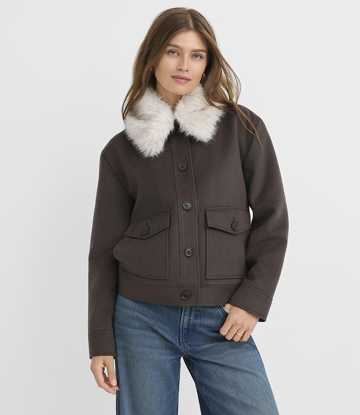 A woman in a brown jacket with a fluffy white collar and blue jeans stands against a plain white background.